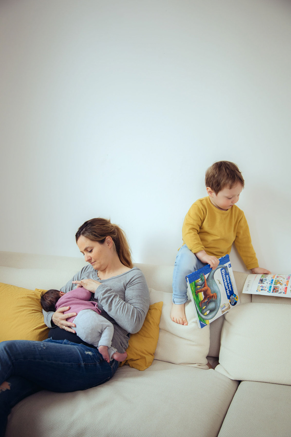 Mutter stillt Baby auf Couch, großer Sohn klettert mit Büchern auf Rückenlehne